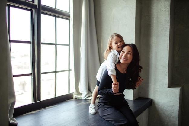 Happy mother and daughter having fun and hugging at home In a dark room sitting on a large window