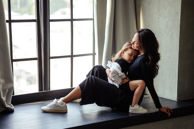 Happy mother and daughter having fun and hugging at home In a dark room sitting on a large window