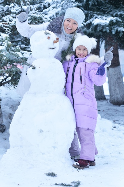 Happy mother and daughter making snowman, showing thumbs up outdoors in winter