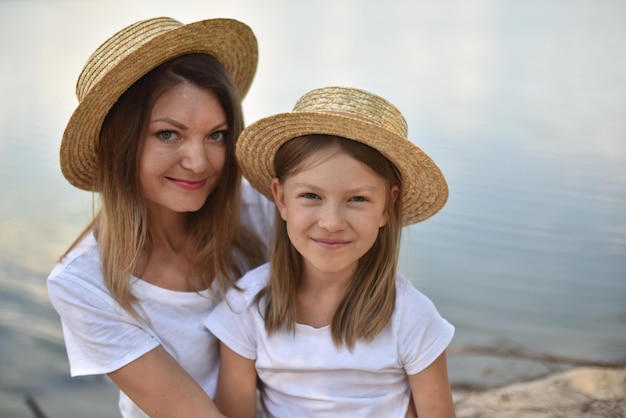 Happy mother and daughter in straw hats and white tshirts look at the camera and sit on river bank