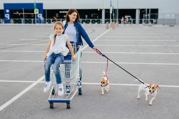 Happy mother daughter and their pets return from shopping centre carry cart pose outdoor have delighted expressions enjoy togetherness have friendly relationships buy something Family concept