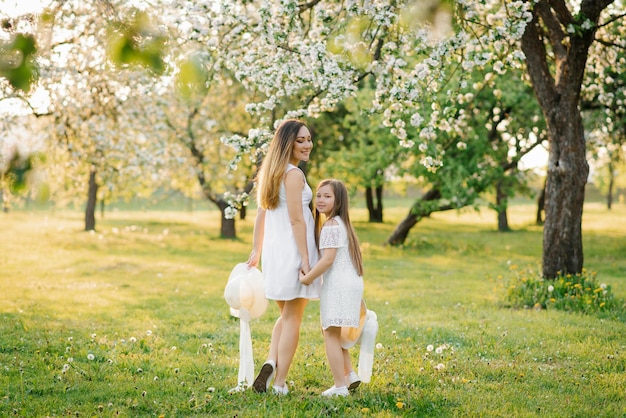 Happy mother and her daughter in white dresses are walking in the spring garden at sunset