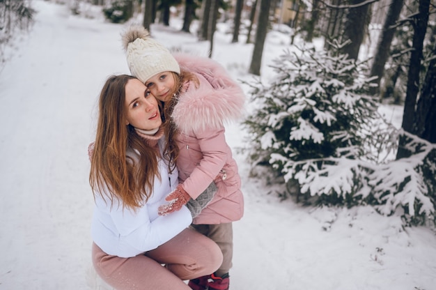 Happy mother and little cute girl in pink warm outwear walking having fun and hugging in snowy white cold winter coniferous forest with spruce woods outdoors