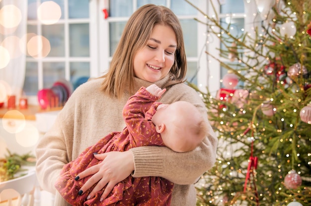 A happy mother plays with a baby in her arms at home not against the background of a decorated room and a Christmas tree