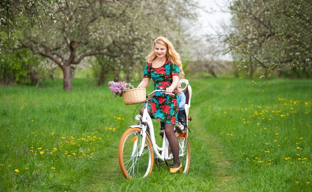 Happy mother riding city bicycle with baby in bicycle chair, in the basket lay a bouquet of lilacs