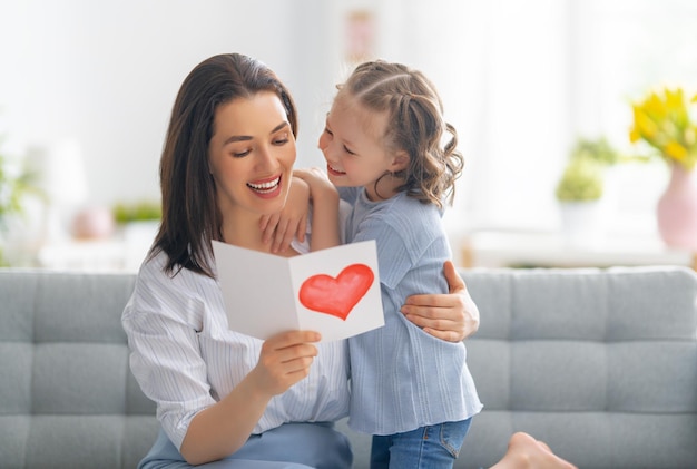 Happy mother's day! Child daughter is congratulating mom and giving her postcard. Mum and girl smiling and hugging. Family holiday and togetherness.