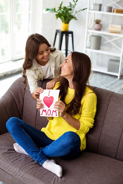 Happy mother's day. Cute daughter congratulates her mom and gives her a postcard. Mum and girl smiling and hugging on the couch. Family holiday and togetherness