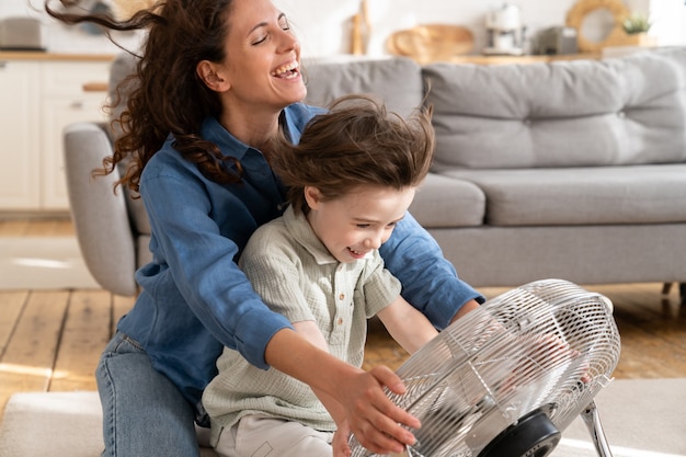 Happy mother and small son enjoy cooling wind blow from ventilator fan sit on floor in living room