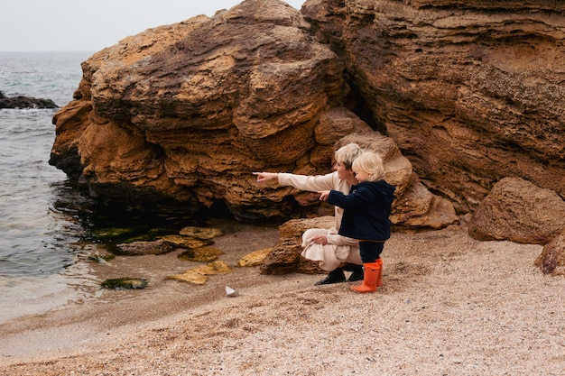 Happy mother and son playing with paper boat on beach near sea in autumn beach