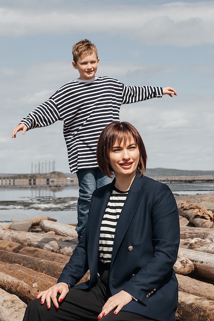 Happy mother and son on the river bank stand on logs in striped vests.