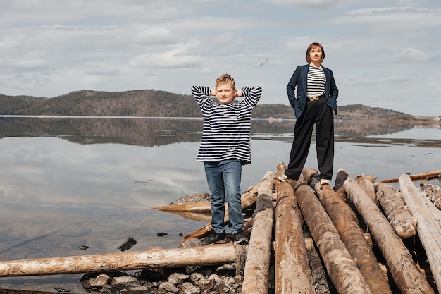 Happy mother and son on the river bank stand on logs in striped vests.