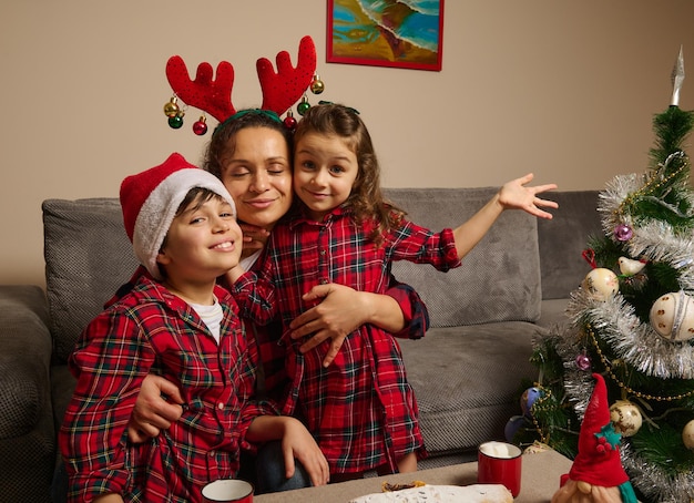 Happy mother with antler hoop hugs her adorable children dressed in red and green plaid clothes and Santa hat on her son's head, celebrating christmas party at home in family circle