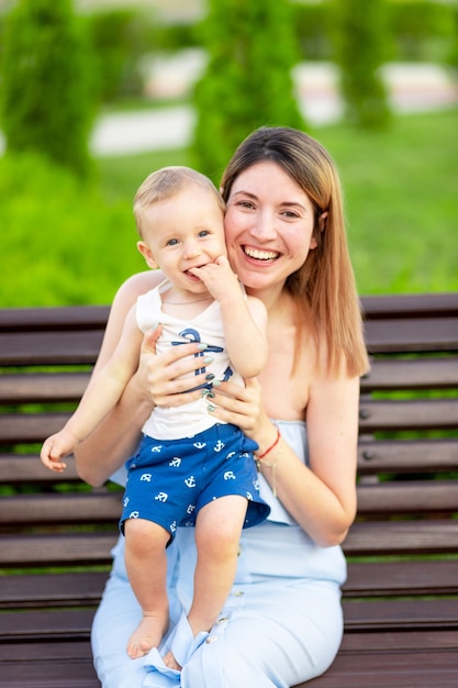 A happy mother with a baby son in the park is sitting on a bench outside in the open air and having fun with a baby in her arms