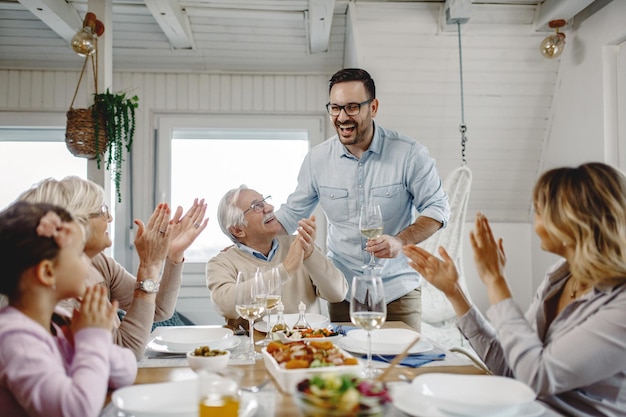 Happy multigeneration family applauding to mid adult man who is proposing a toast during lunch at dining table