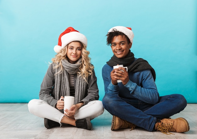 Happy multiracial teenage couple sitting isolated over blue wall, wearing christmas hats, holding cups