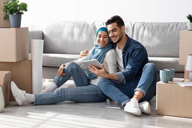 Happy muslim family sitting on floor in new apartment with digital tablet among boxes with belongings, buying furniture for house or working on design together, drinking tea and having conversation