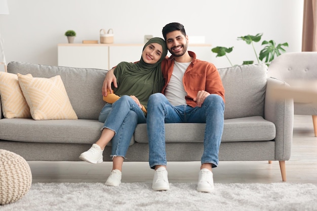 Happy muslim spouses sitting on sofa embracing posing at home