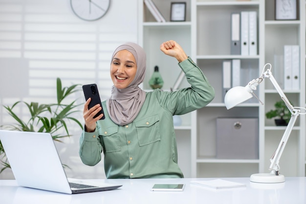 Happy muslim woman in hijab with raised fist in victory gesture using smartphone and laptop in a