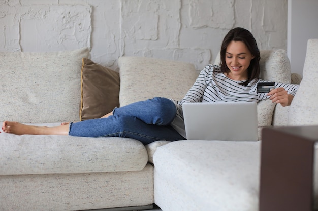 Happy natural brunette using credit card and laptop in the living room.