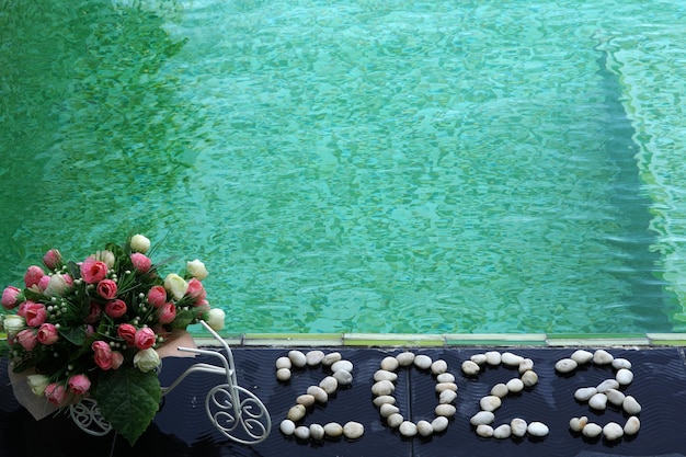 Happy New Year 2023 written by stones with artificial roses flowers and green swimming pool.