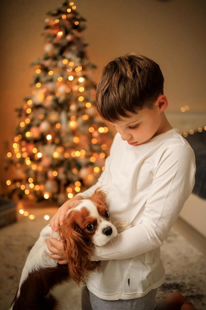 Happy New Year a boy in light homemade pajamas hugs his pet cavalier king charles spaniel at home in the bedroom near the Christmas tree