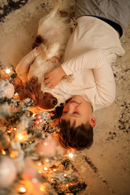 Happy New Year a boy in light homemade pajamas hugs his pet cavalier king charles spaniel at home in the bedroom near the Christmas tree