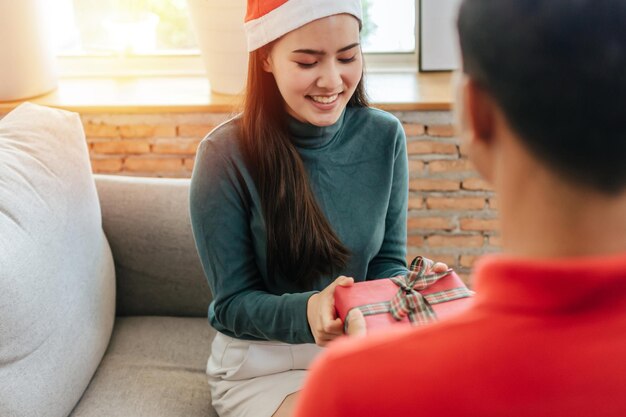 Happy new year. lovely romantic young couple surprising and giving christmas gift box to his girlfriend sitting on sofa christmas celebrating in house, holiday and merry christmas festival concept