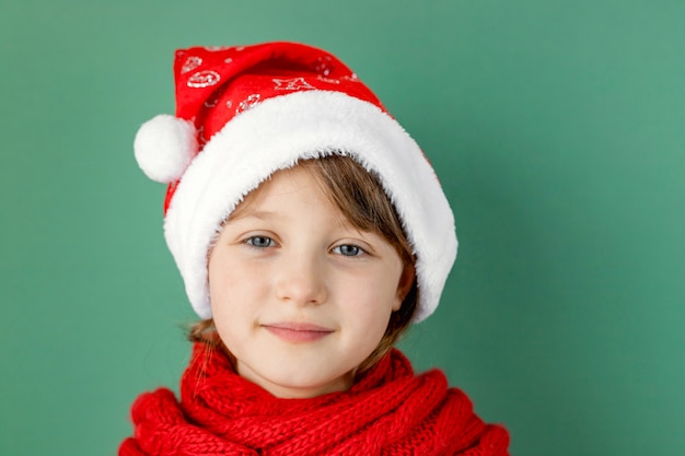 Happy new year and merry christmas. . Cute little caucasian girl in a santa hat and a red scarf on a green wall.