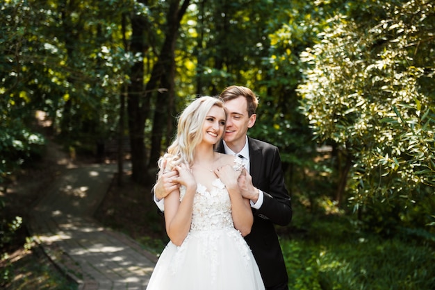 Happy Newlywed woman and man embracing and kissing in green park. Loving wedding couple outdoor. Bride and groom. Bride and groom at wedding day walking outdoors on summer green nature