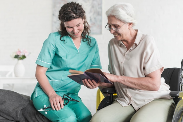 Happy nurse looking at book hold by senior female patient sitting on wheelchair