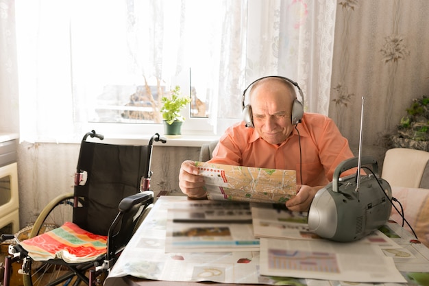 Happy Old Bald Man, Sitting at the Living Room, Reading Newspaper While Listening at Radio with Headset