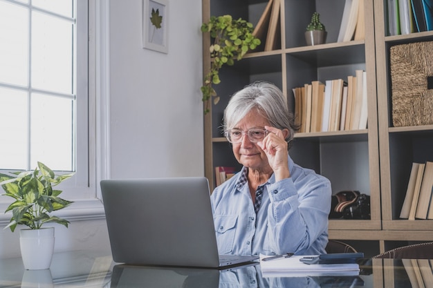 Happy old caucasian businesswoman smiling working online watching webinar podcast on laptop and learning education course conference calling make notes sit at work desk, elearning concept