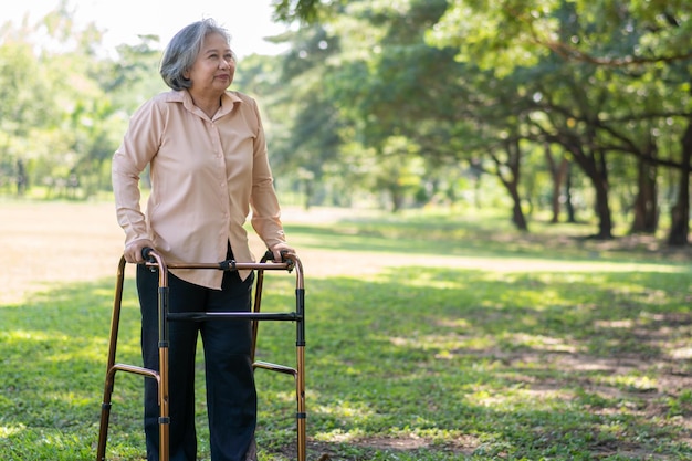 Happy old elderly Asian woman uses a walker for osteoarthritis rehabilitation physiotherapy in park Concept of happy retirement With care from a caregiver and Savings and senior health insurance