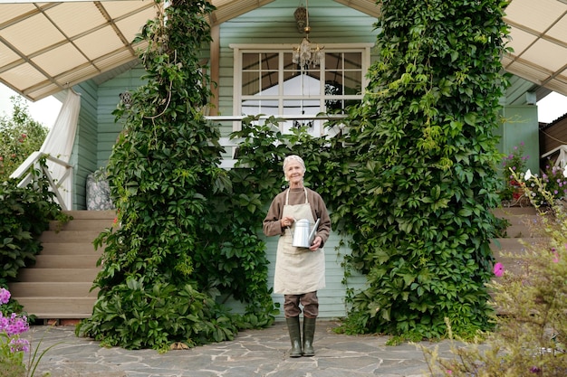 Happy old woman in workwear standing in front of camera against cottage