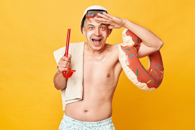 Happy overjoyed tourist man at summer resort standing with rubber ring isolated over yellow background looking far with hand near forehead laughing happily