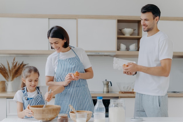 Happy parents cook together with daughter pose at modern home kitchen give eggs to add to dough small kid whisks ingredients in bowl have glad expressions Food and family concept Cooking breakfast