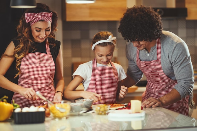 Happy parents and their daughter are preparing meal together in the kitchen while little girl is putting ketchup on the pizza dough.