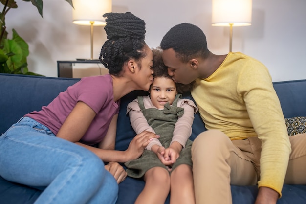 Happy people. Dark skinned young adult mom and dad kissing their little cute daughter sitting between parents on sofa at home