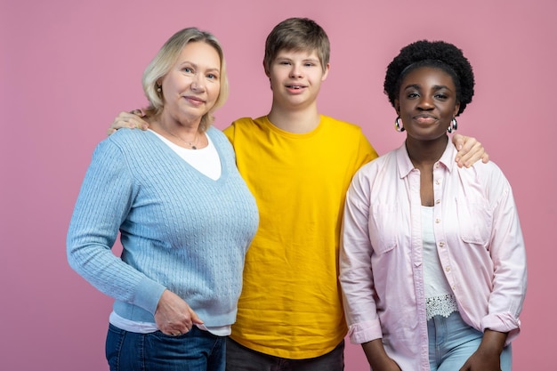 Happy people. Guy with down syndrome hugging mom and dark-skinned woman standing smiling looking at camera on light background