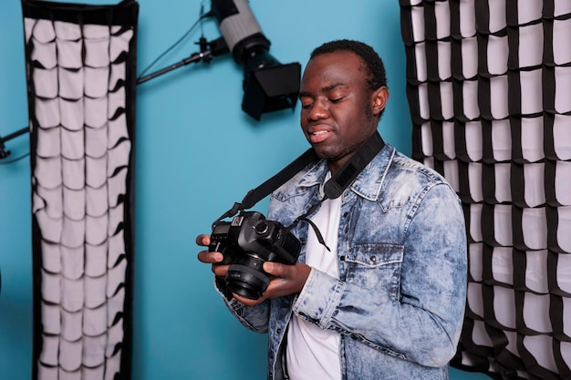 Happy photography enthusiast with DSLR device standing on blue background. Smiling photographer checking camera gallery while standing in production studio equipped with softboxes and spotlights.