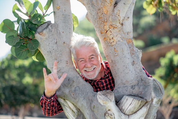 Happy playful senior bearded man partially hidden by a tree trunk in the park looking at camera