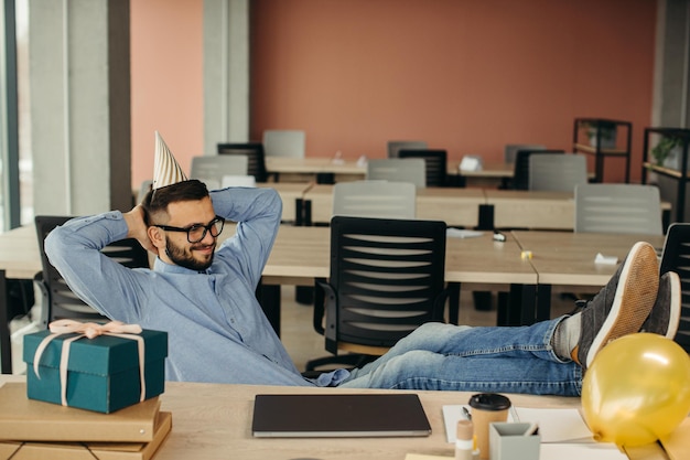 Happy positive bearded office worker in cone hat sitting at workplace with laptop and gift with festive ribbon