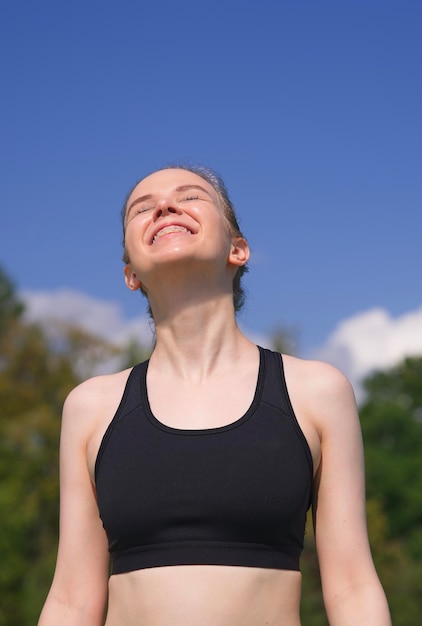 Happy positive fit fitness girl joyful young woman is enjoying sun good summer weather sunbathing at sunny day on natural green and sky background Vertical photo