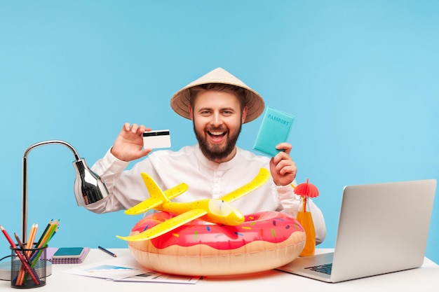 Happy positive man sitting at workplace with rubber ring and toy plane holding passport and credit card planning vacations abroad booking tickets Indoor studio shot isolated on blue background