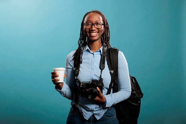 Happy positive photography entusiast having DSLR photo device and cup of coffee while standing on blue background. Young adult woman with photographing hobby smiling at camera.