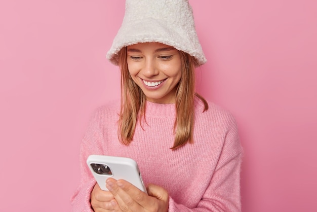 Happy positive woman smiles gladfully uses mobile phone for communication downloads useful application wears white winter panama and sweater isolated over pink background. Modern technologies