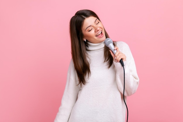 Happy positive young female standing holding microphone singing songs in karaoke having fun alone wearing white casual style sweater Indoor studio shot isolated on pink background