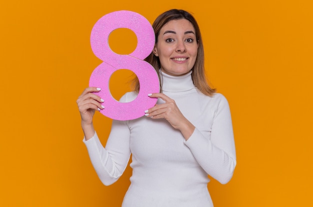 happy and positive young woman in white turtleneck holding number eight