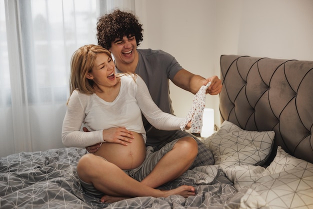 Happy pregnant couple having fun while looking new clothing for upcoming baby at home in the bedroom.