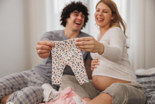 Happy pregnant couple showing baby clothes while relaxing on a bed in bedroom at home.